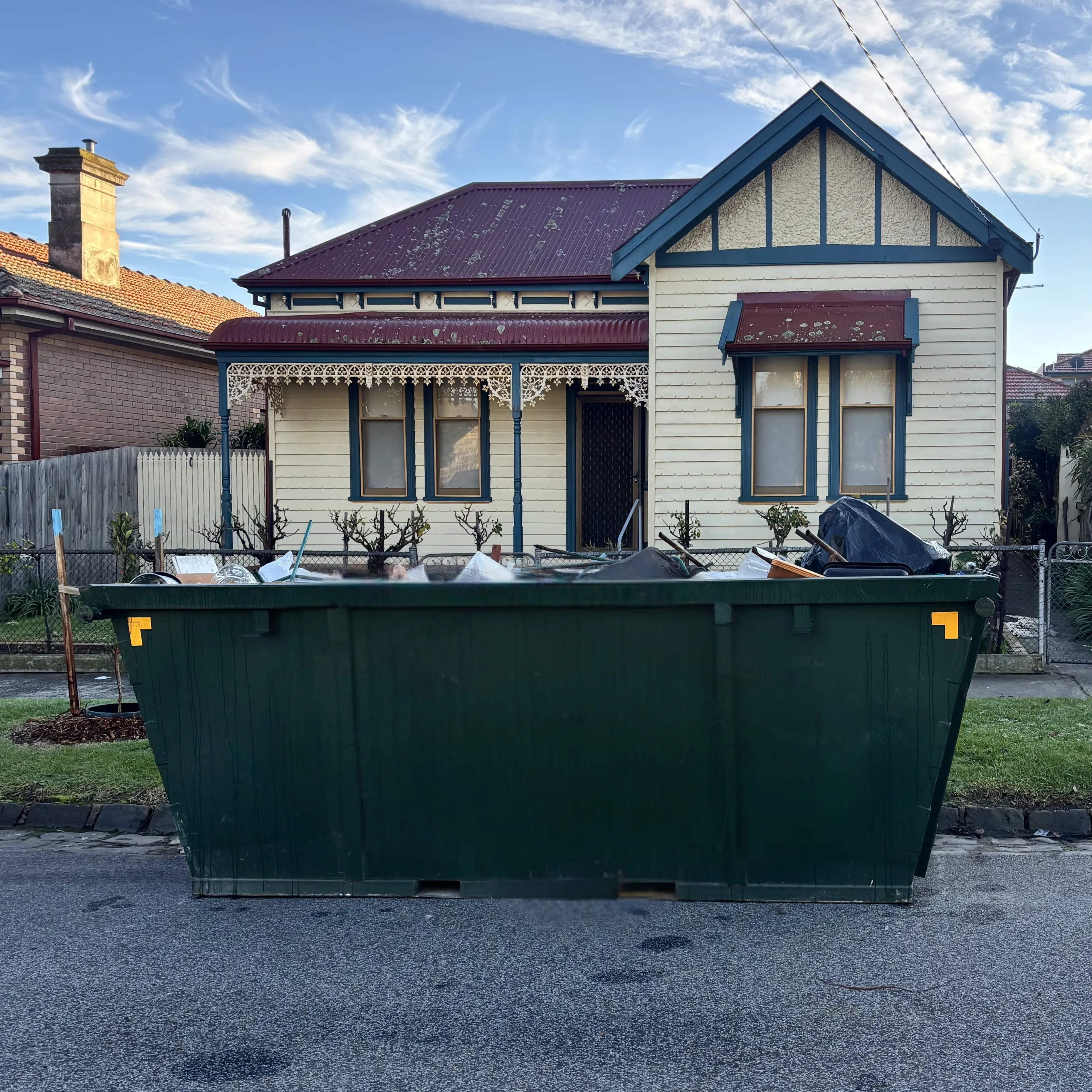 A skip bin on the street outside a weatherboard cottage, filled with household waste